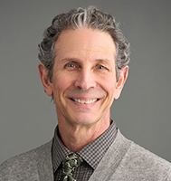 February 2025, man with grey hair, beige sweater with checkered shirt, tie, against a grey background