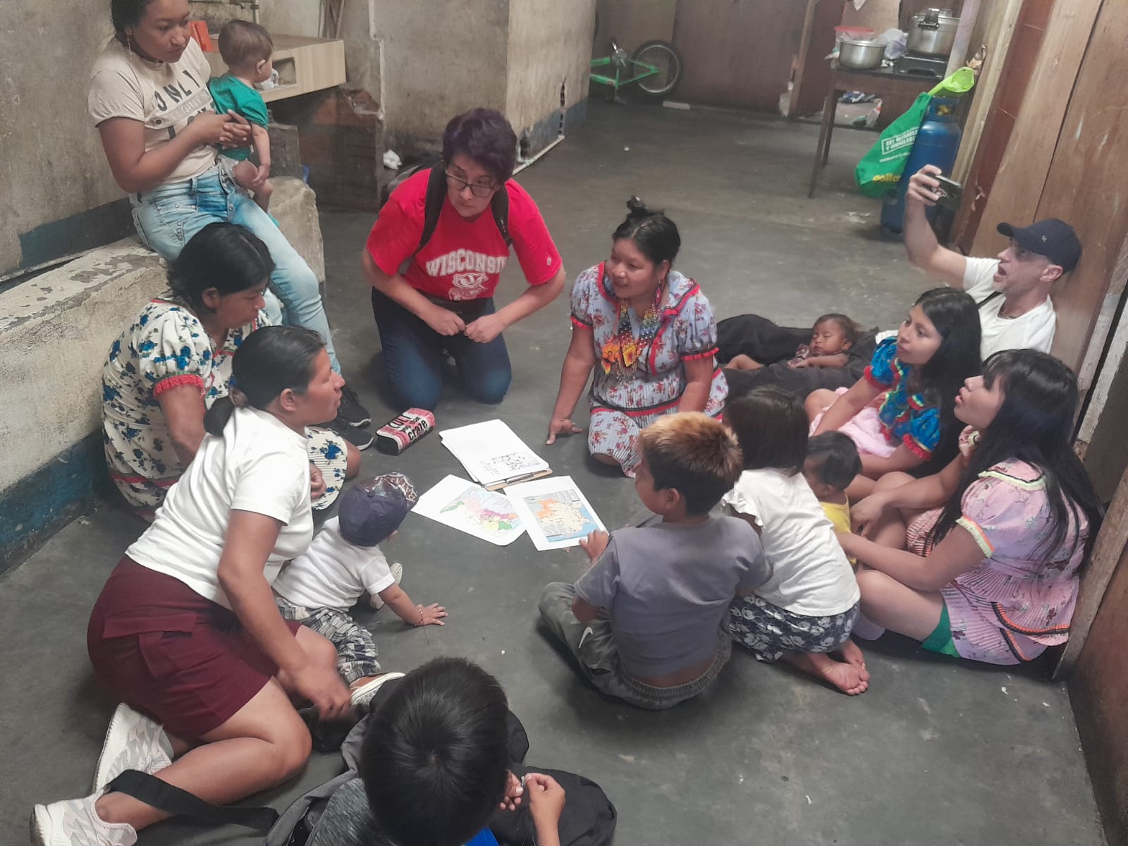 June 2024, woman with dark hair and glasses in a red Wisconsin shirt sitting on the floor with children and women
