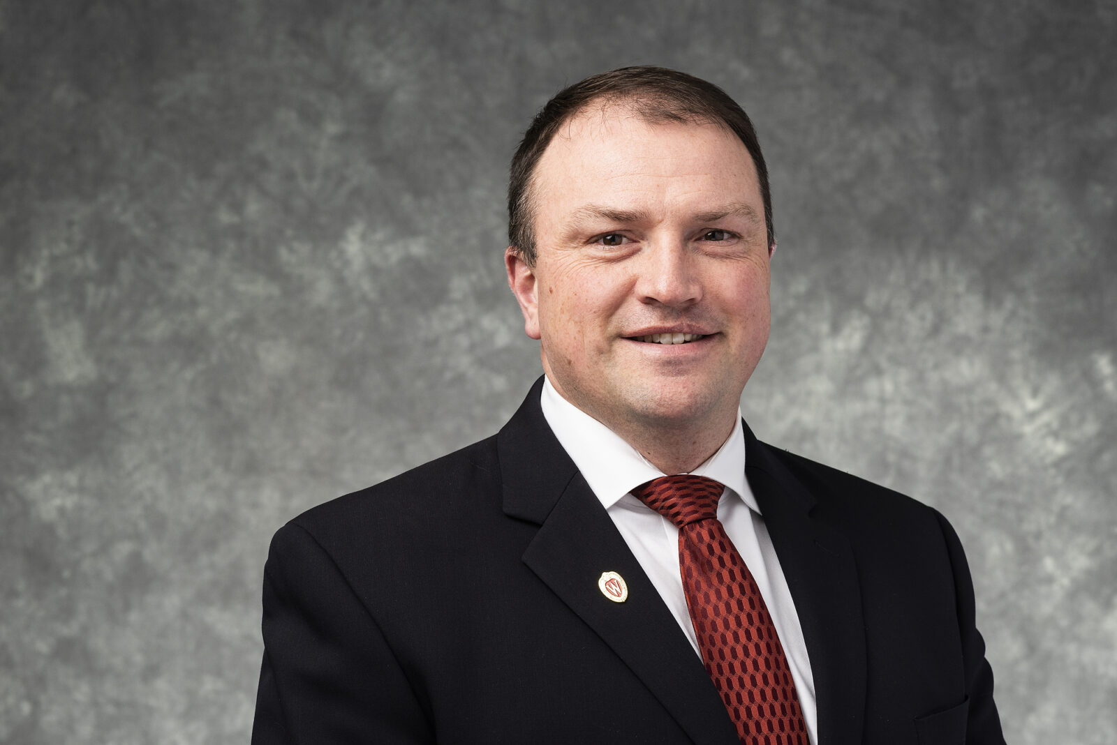 September 2024, man with dark hair, dark jacket, white shirt, red tie against a grey background