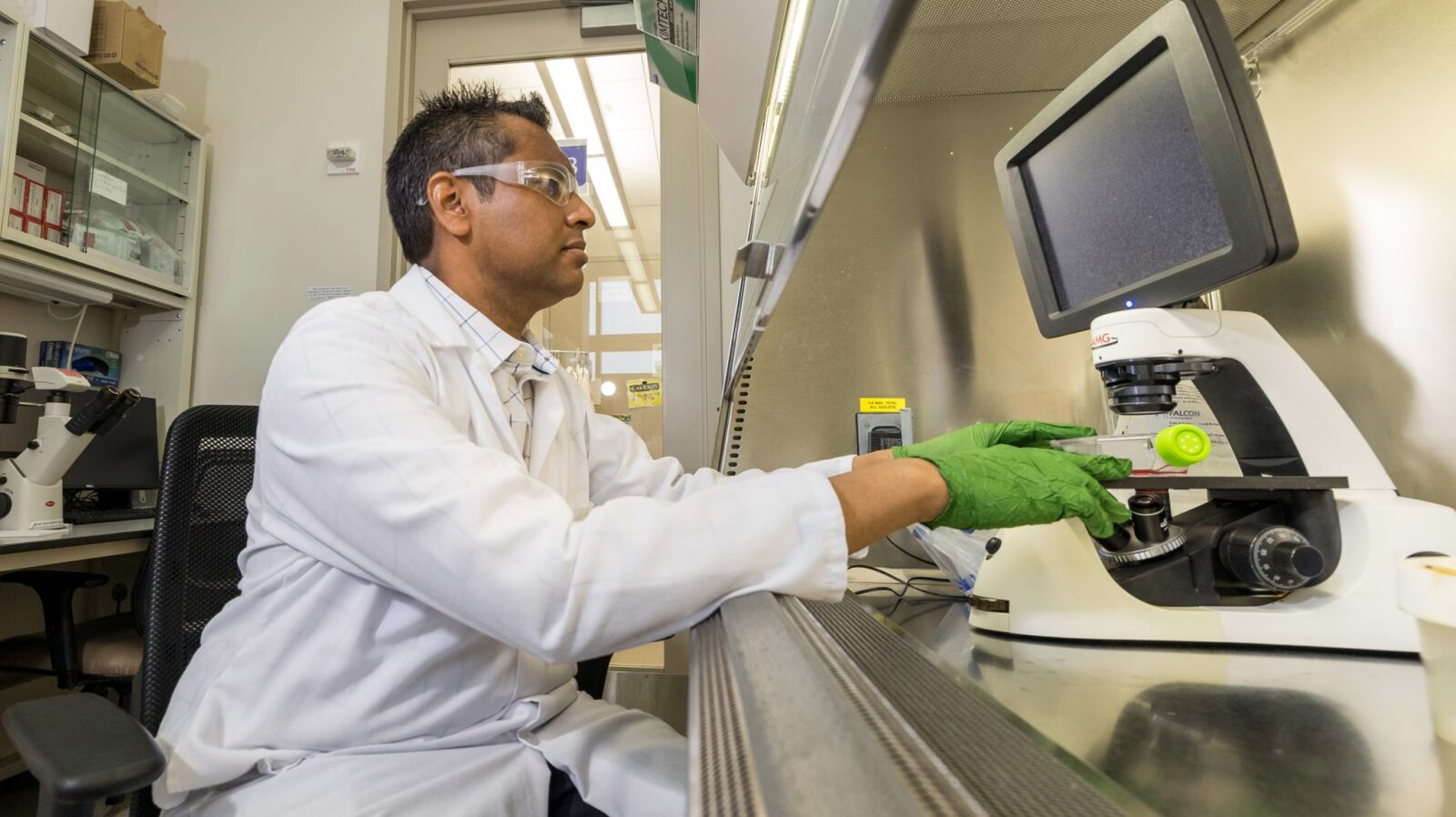 March 2025; Kris Saha in his lab at UW; a man wearing a white lab coat, goggles, and green gloves working with a microscope