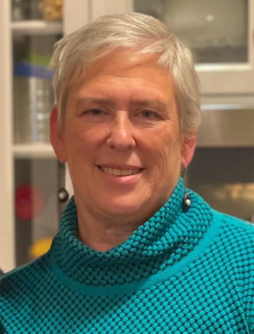 February 2025, woman with white hair, long earrings, green turtleneck against a bookshelf backgroun