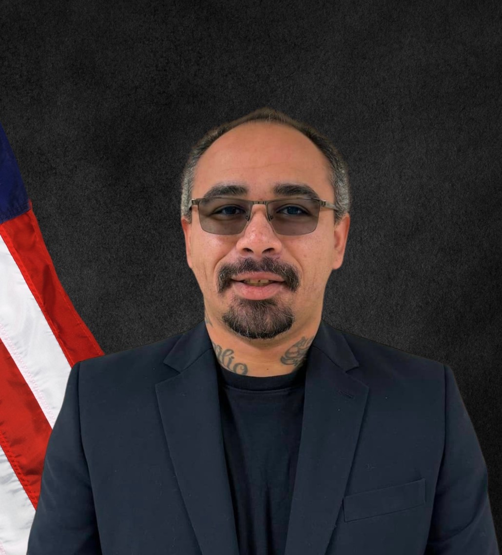 December 2024; man with dark hair, glasses, dark suit, with an American flag against dark background