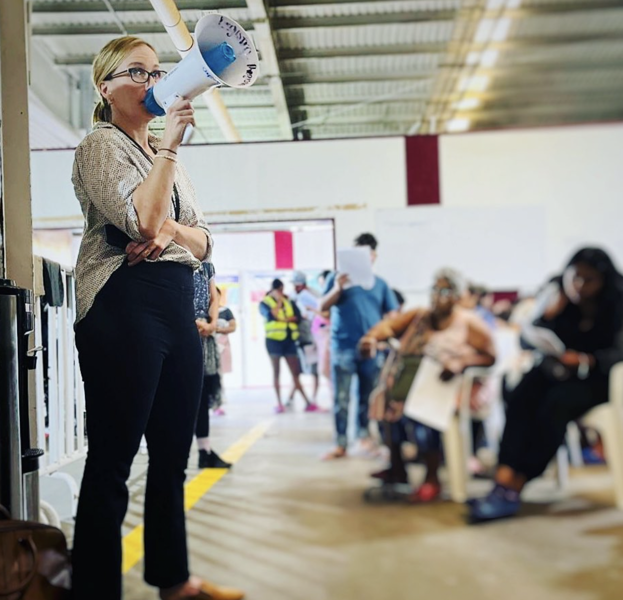 March 2024, a woman with light hair and glasses speaking into a megaphone to a group of people sitting and walking in a room with a brown floor and white walls