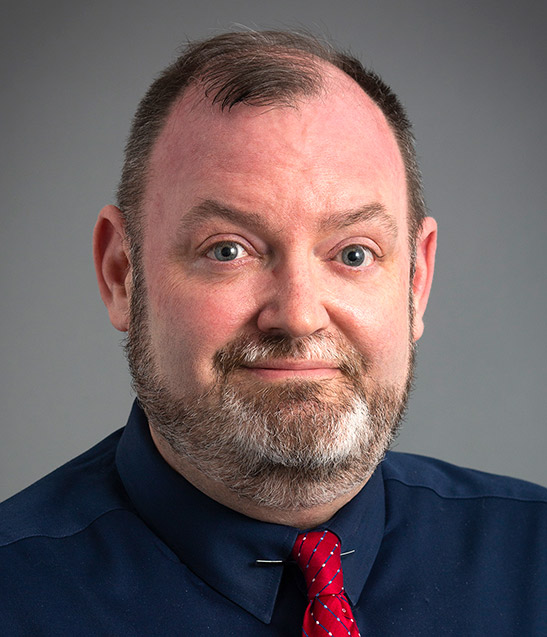 November 2024, man with dark hair, grey beard, dark shirt, red tie against a grey background