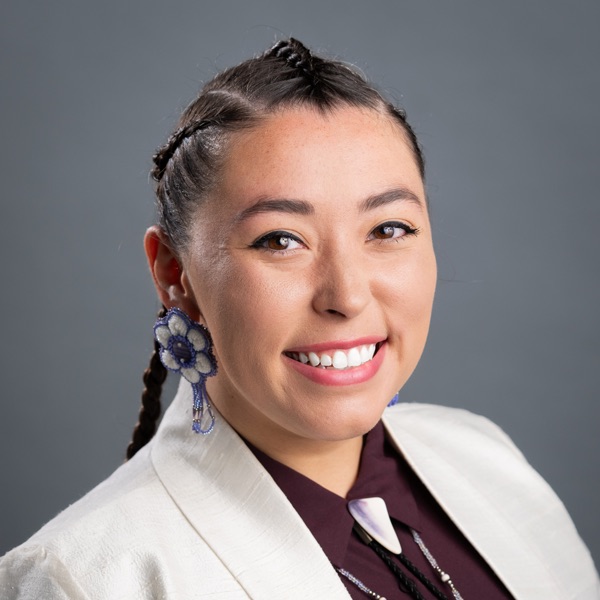 December 2024; woman with dark braided hair, white suit with maroon shirt, silver tie against a grey background