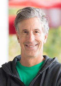 February 2024, man with grey hair, dark hoodie, green shirt against an blurred outdoor background