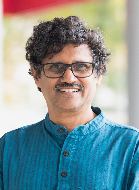 Giri Venkataramanan November 2023, man with dark hair and mustache, blue shirt against a light filled window