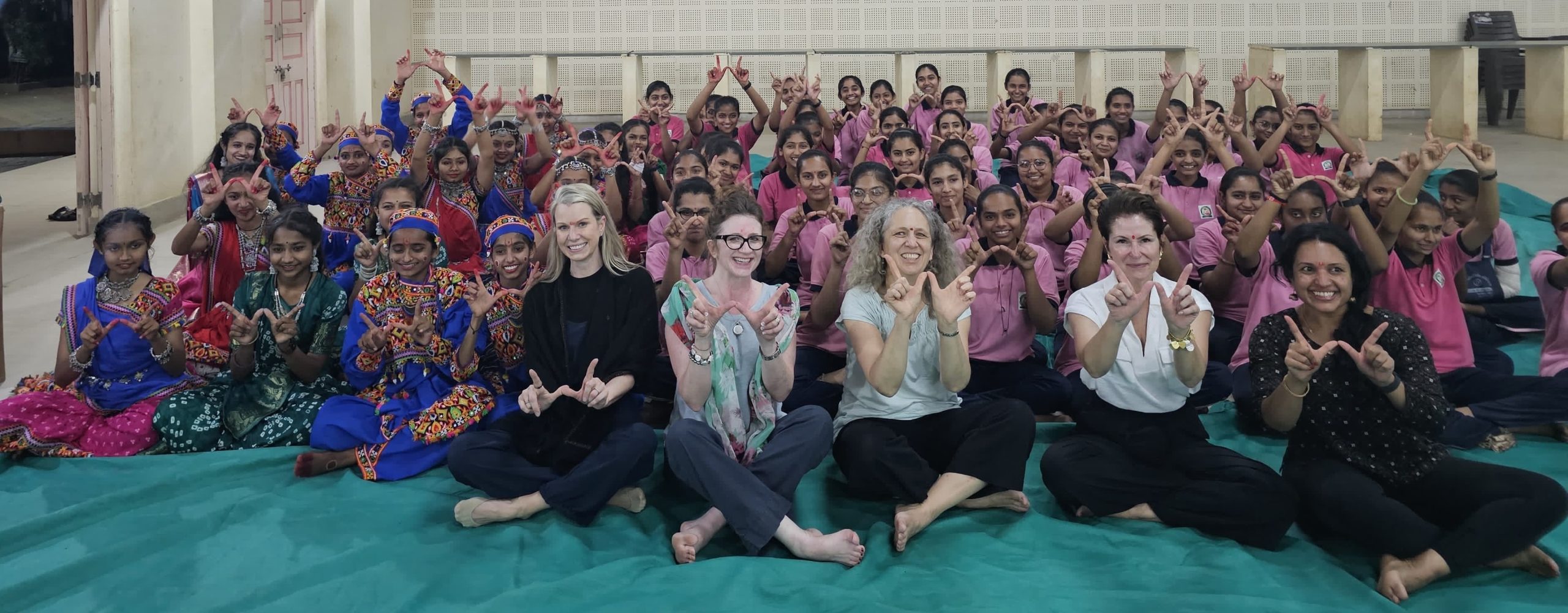 FIVE WOMEN SITTING CROSS LEGGED IN FRONT OF A GROUP OF SCHOOL-AGED GIRLS IN INDIA; SOME OF THE GIRLS ARE WEARING PINK SHIRTS AND OTHERS ARE WEARING COLORFUL SARIS. ALL ARE HOLDING UP THE UW "W" WITH THEIR HANDS