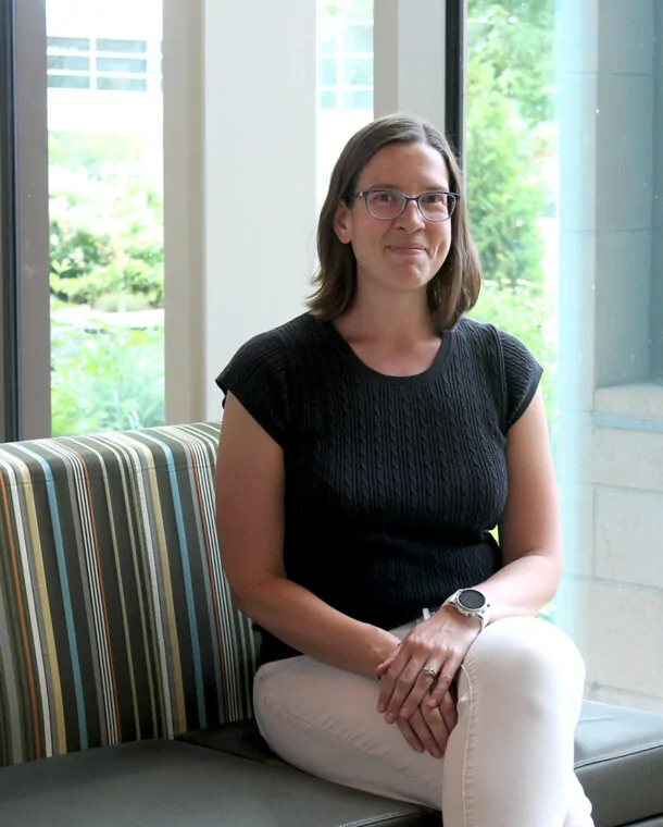 photo of Heather Barkholtz; woman sitting on a striped couch with dark hair, glasses, dark top and light pants