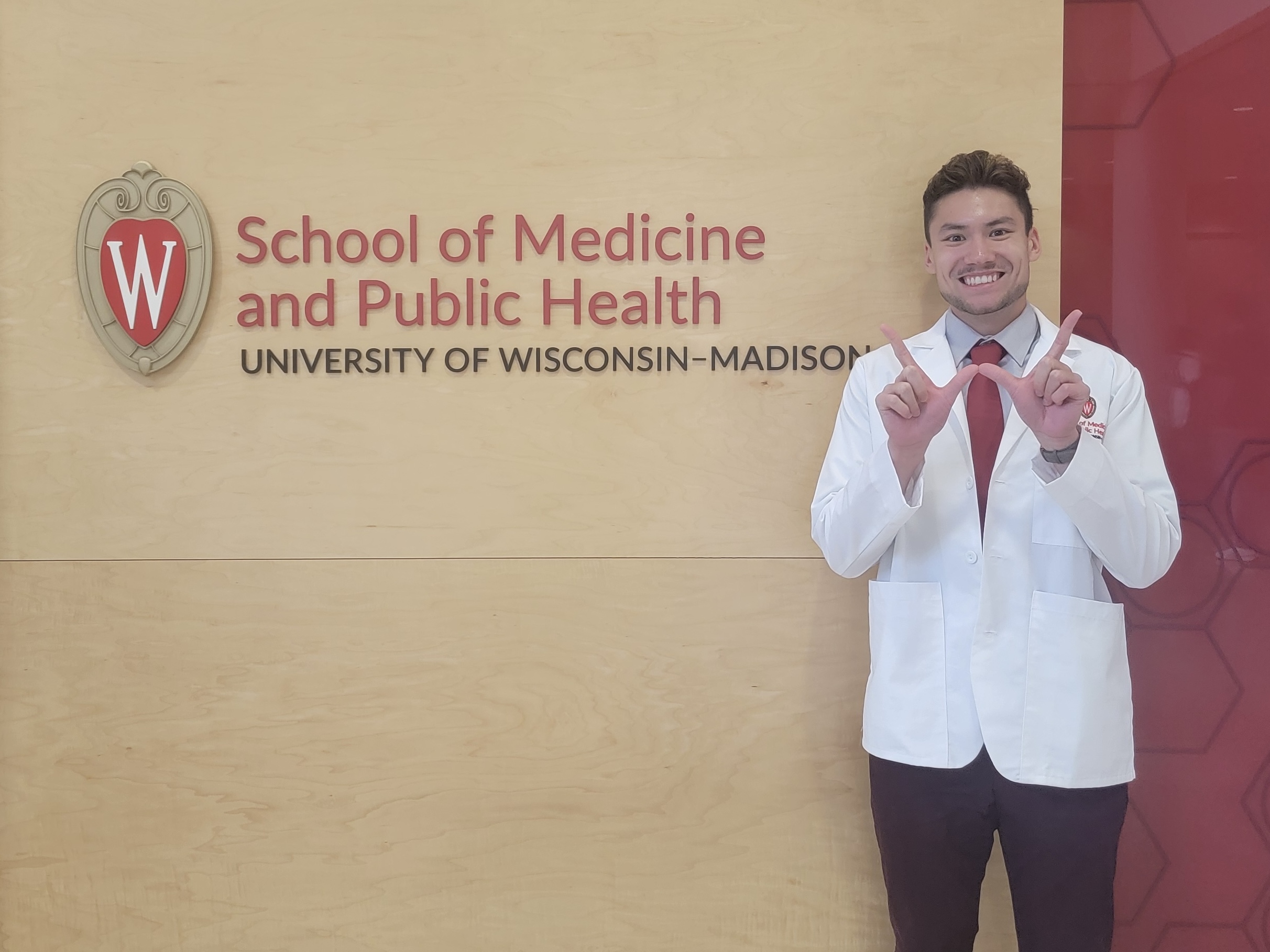 Jonathan Bryan in a white lab coat with a red tie, holding hands in a W formation in front of a sign for the UW School of Medicine and Public Health