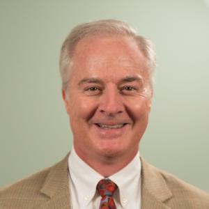 headshot of James Conway; man with grey hair, white shirt, tan jacket against a beige background