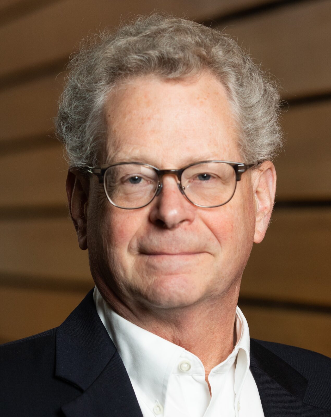 Craig Hedberg headshot, man with grey hair, glasses, white shirt and dark jacket against a brown and black stripe background