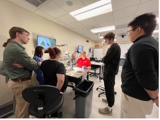man in red sitting in a lab setting teaching with six students standing and one other man sitting next to the man in red