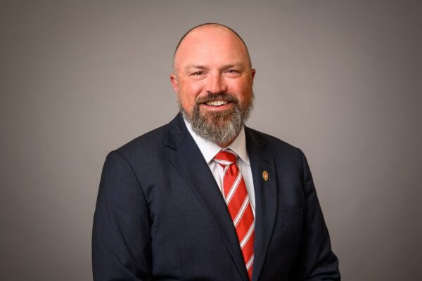 headshot of Keith Poulsen; bald man with beard, white shirt, dark jacket, red & white tie against a dark background
