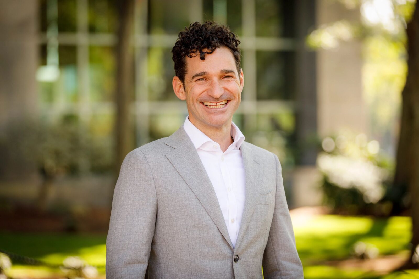 headshot of Craig Spencer, man with dark hair, white shirt, grey suit in an outdoor setting with a class building in the background