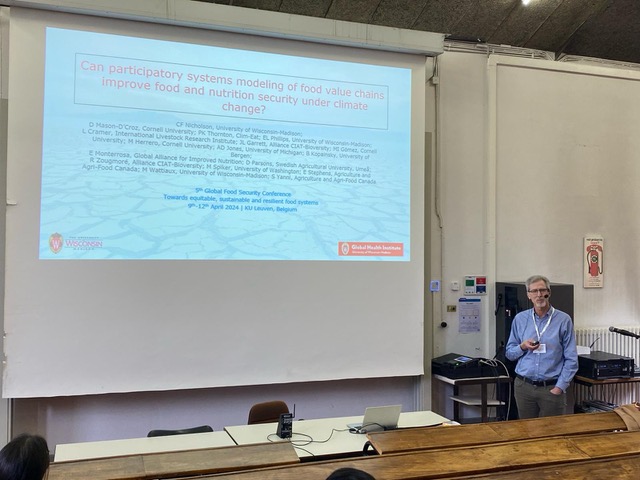 Dr Nicholson presenting work on the workshop takeaways; man with grey hair, glasses, white lanyard, and blue shirt standing next to a large white projection screen with text