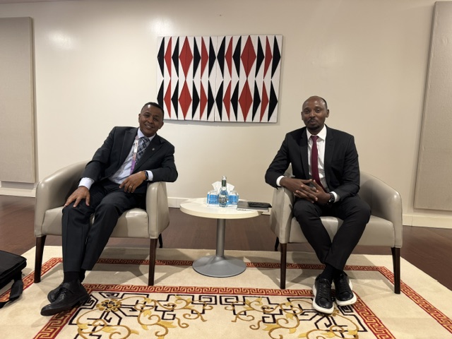 two men with dark hair and suits sitting on grey chairs with a white wall and red & black artwork on teh wall and red, black, and grey carpet
