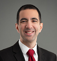 man with dark hair, wearing a white shirt, red tie and dark jacket against a grey background