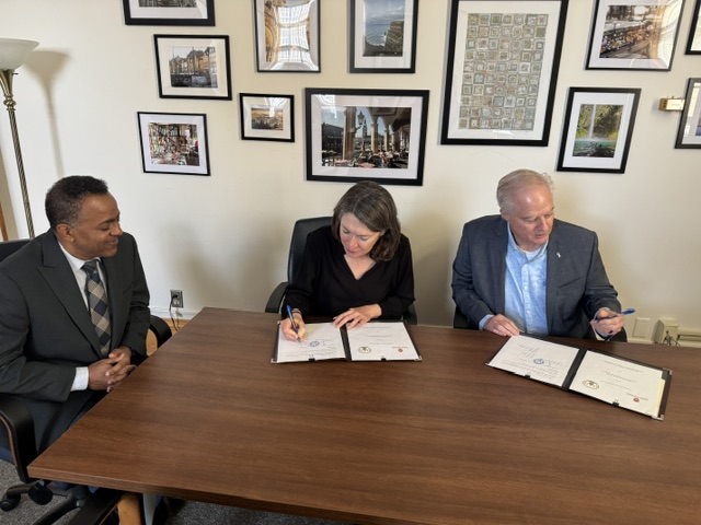 three people sitting at a table, two on the right have pens in hands signing documents with framed photos on the wall in the background