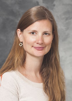 woman with long light brown hair, diamond-shaped earring, wearing a light top against a grey background