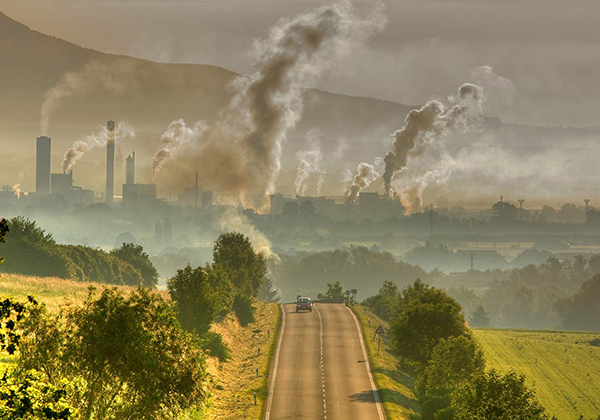 Smoke rises from factories in the background of a car driving through fields and trees.