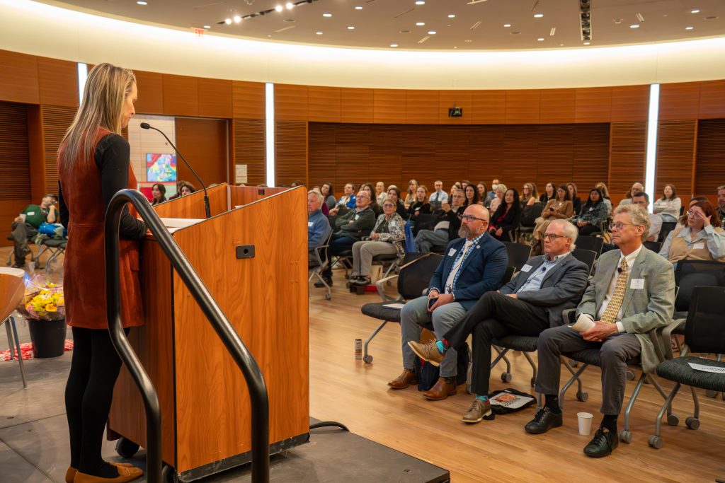 Photo from 2026 Global Health Symposium at the Discovery Building; Calyn Ostrowski of GHI at the lecturn facing the crowd with members looking on
