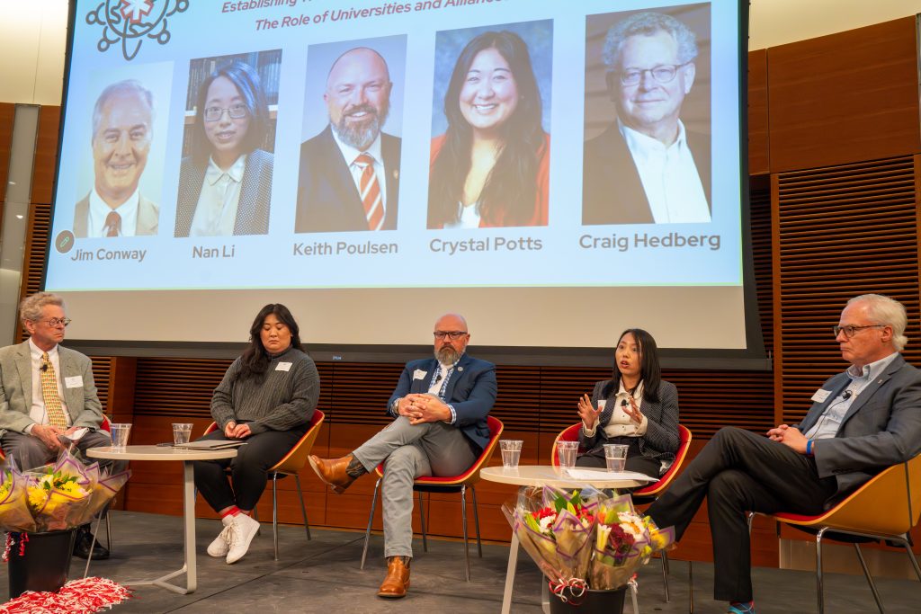 Photo from 2026 Global Health Symposium at the Discovery Building; five panelists sitting on stage with a screen with their five photos in the background