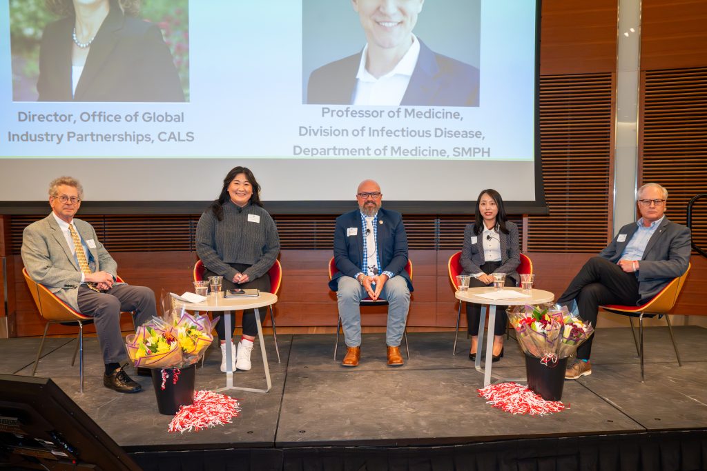Photo from 2026 Global Health Symposium at the Discovery Building; five panelists sitting on stage with flowers and red & white decor with screen in the background