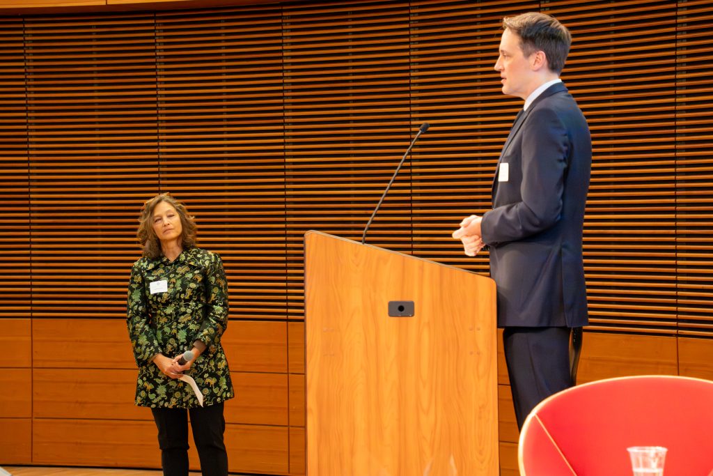 Photo from 2026 Global Health Symposium at the Discovery Building; Dr. Ryan Westergaard at the lecturn with Dr. Jennifer Kushner standing below with a green decorative jacket