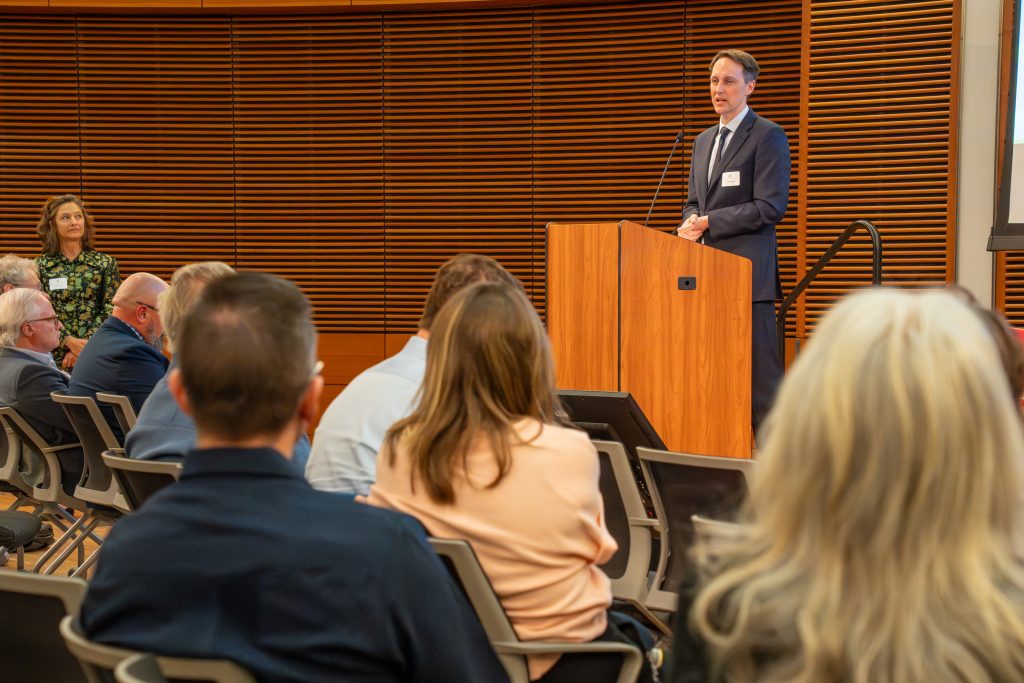 Photo from 2026 Global Health Symposium at the Discovery Building; Dr. Westergaard at lecturn with backs of crowd members sitting