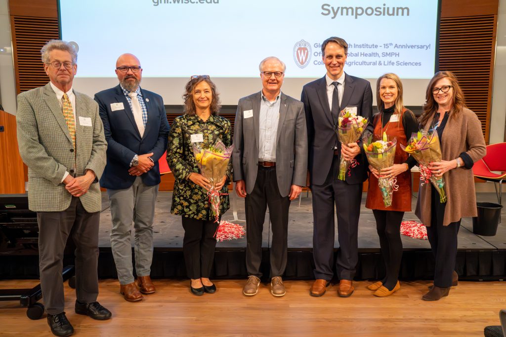 Photo from 2026 Global Health Symposium at the Discovery Building; seven people standing in front of a stage, four holding bouquets of flowers