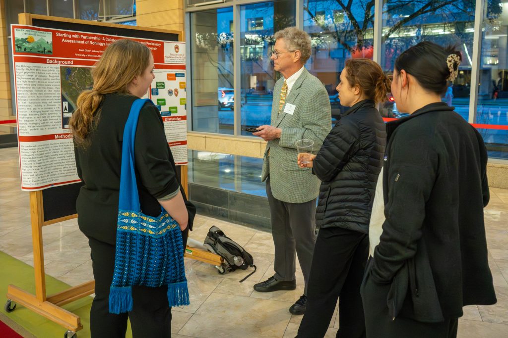 Photo from 2026 Global Health Symposium at the Discovery Building; Dr Hedberg looking at a poster with three students at the discovery building atrium